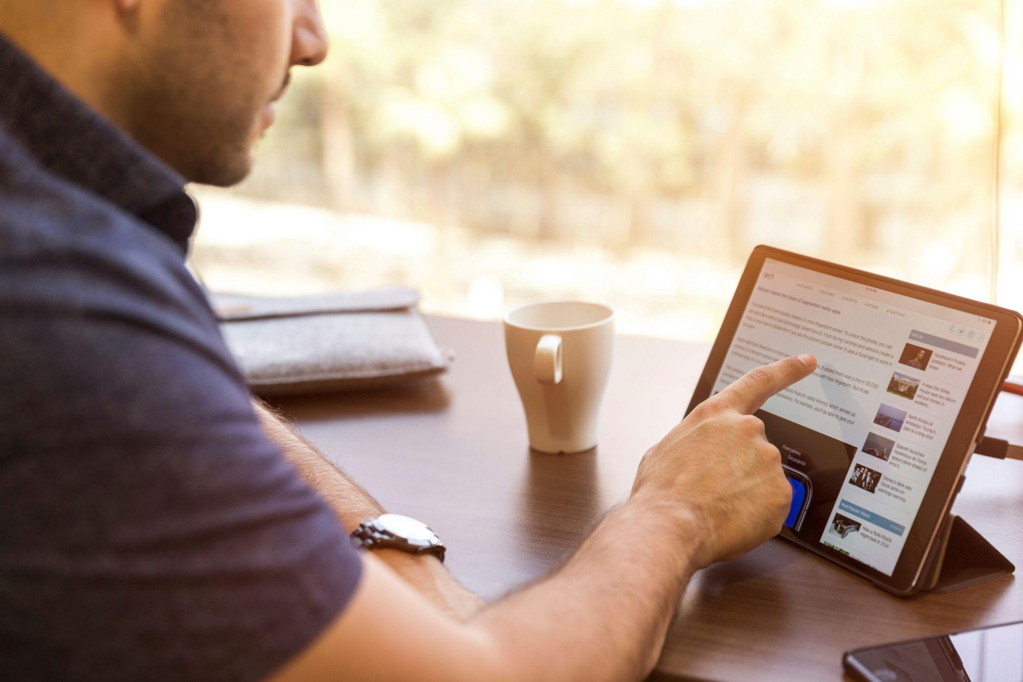 Hombre usando una tablet para leer noticias o artículos en línea mientras trabaja en una mesa con una taza de café.
