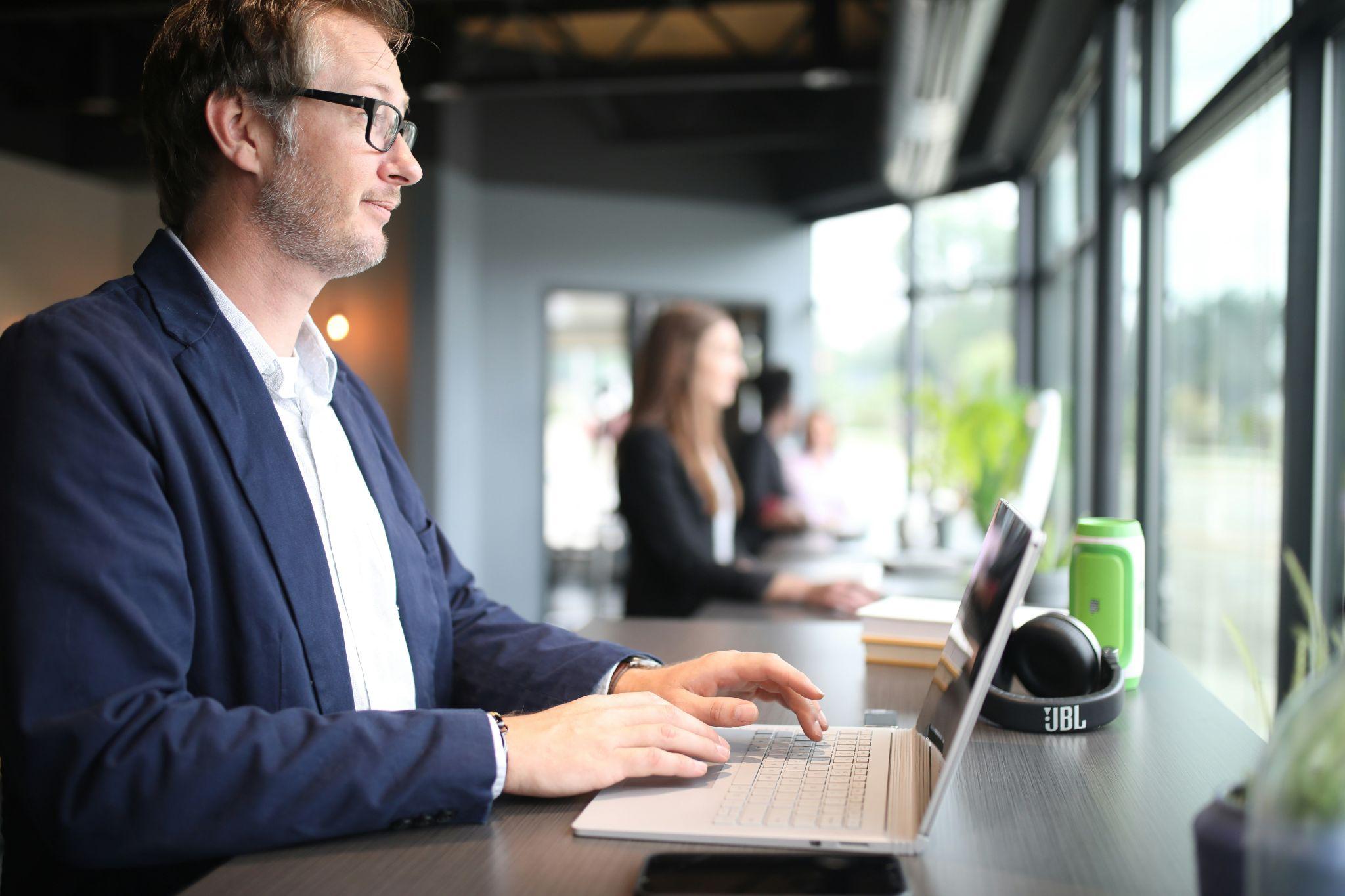 Hombre trabajando en laptop en oficina moderna con ventanales, con otras personas en segundo plano.