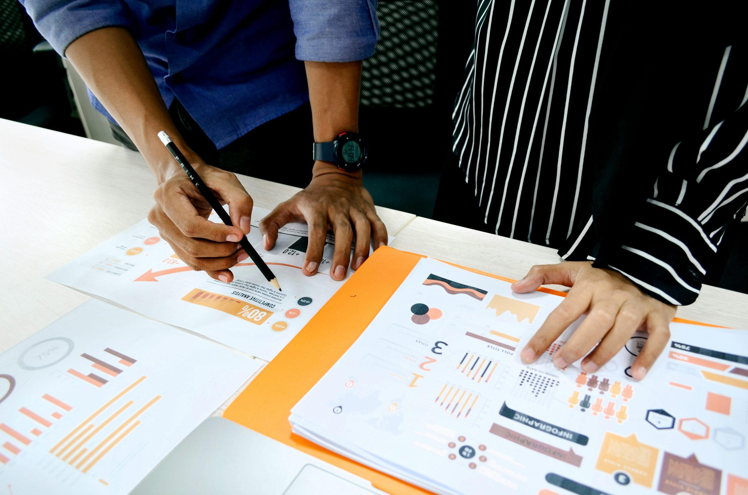 Dos personas colaboran sobre tablas y gráficos en una mesa. Las manos señalan los datos, sugiriendo trabajo en equipo y análisis en un entorno empresarial. Los elementos visuales coloridos crean un ambiente enfocado.