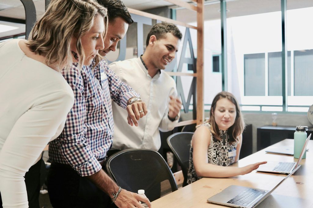 Un grupo de cuatro personas se reúne alrededor de una computadora portátil en una oficina. Tres están de pie, comprometidos y sonrientes, mientras una mujer está sentada señalando la pantalla. La atmósfera es colaborativa y positiva.