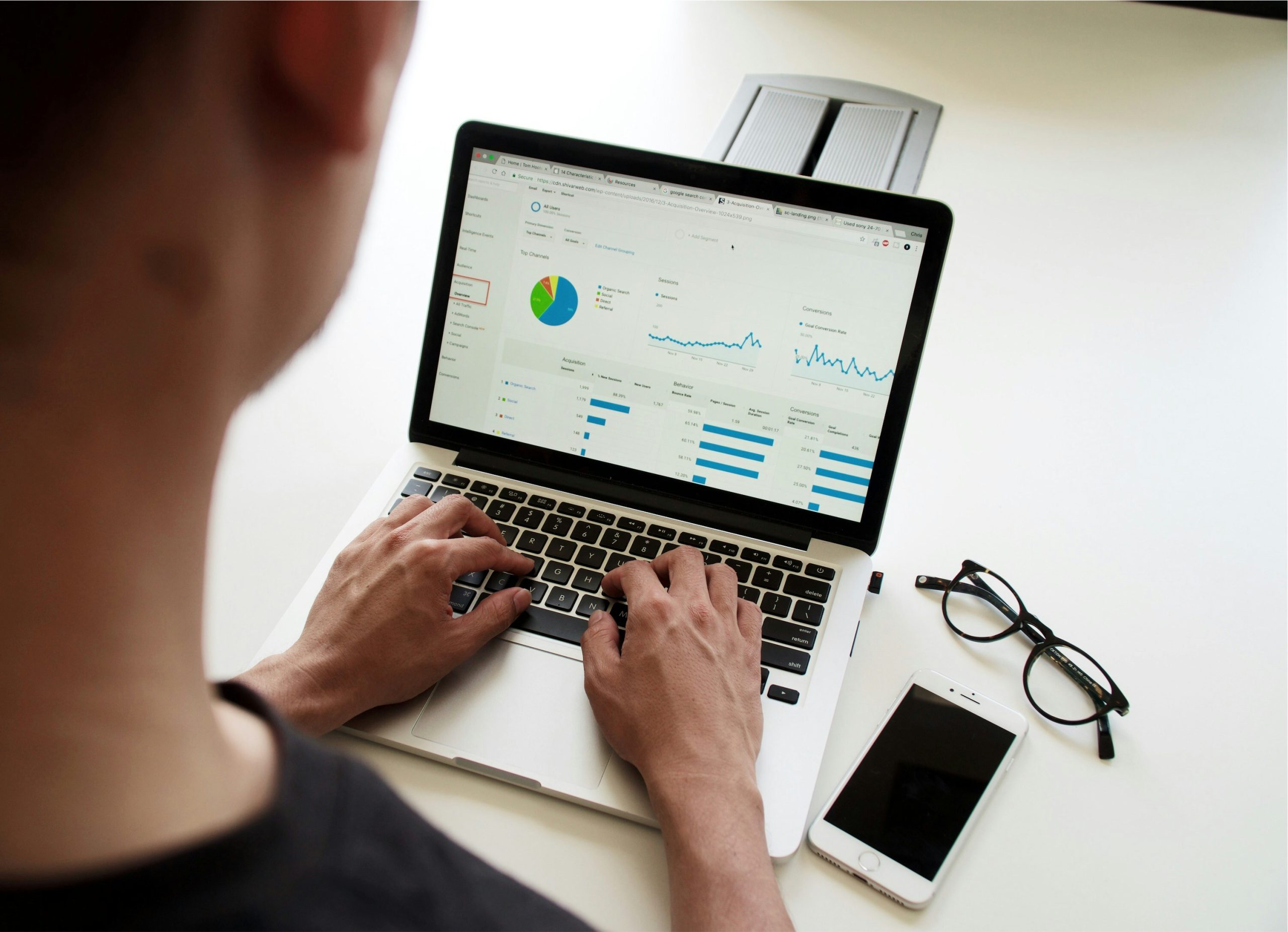 Una persona está usando una computadora portátil que muestra análisis de datos con tablas y gráficos. Cerca, un teléfono inteligente y unas gafas descansan sobre la mesa, transmitiendo un entorno de trabajo enfocado.