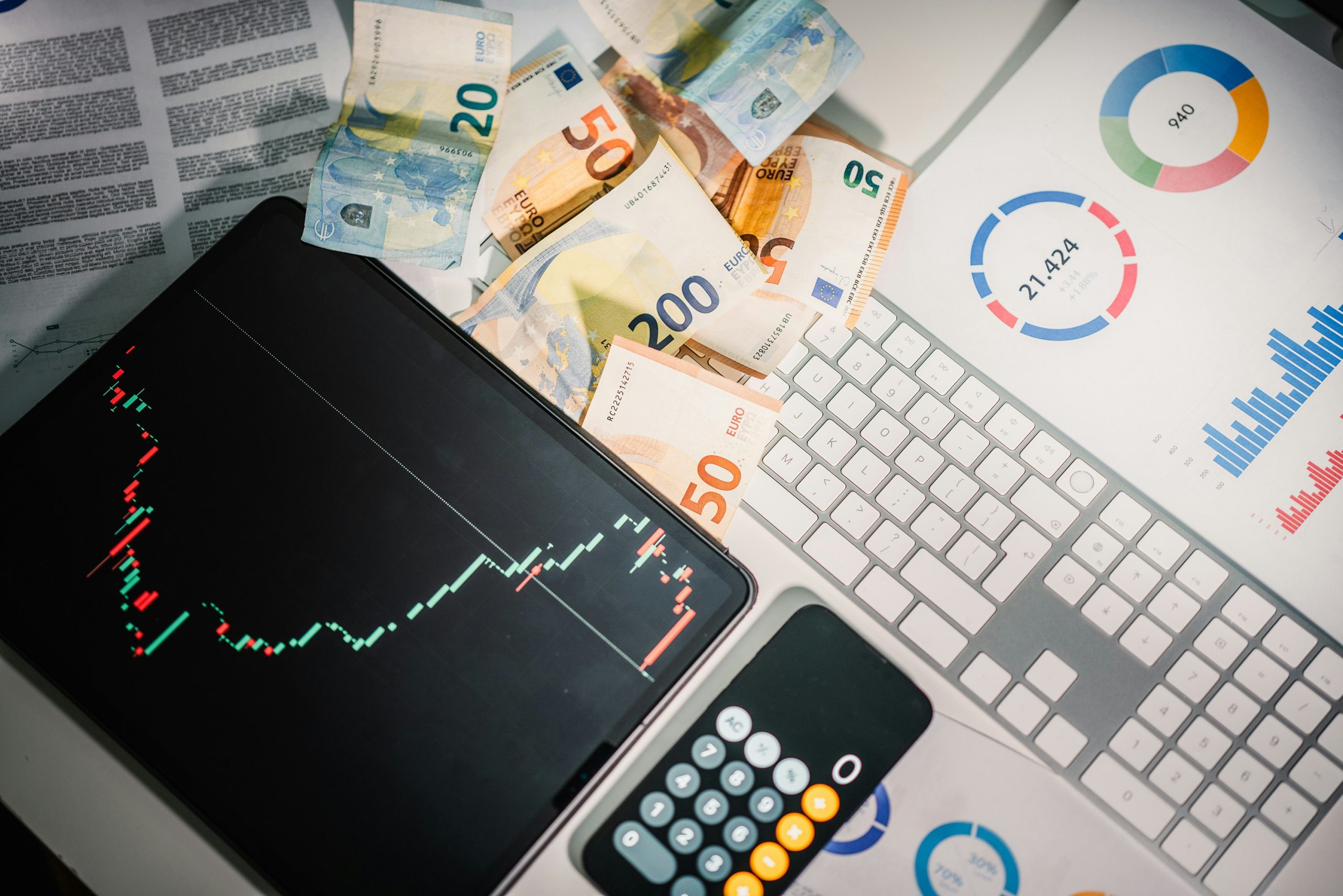 Tableta que muestra un gráfico de acciones, billetes de euro esparcidos, un teclado, una calculadora y gráficos de negocios en papel, transmitiendo un tema de análisis financiero.