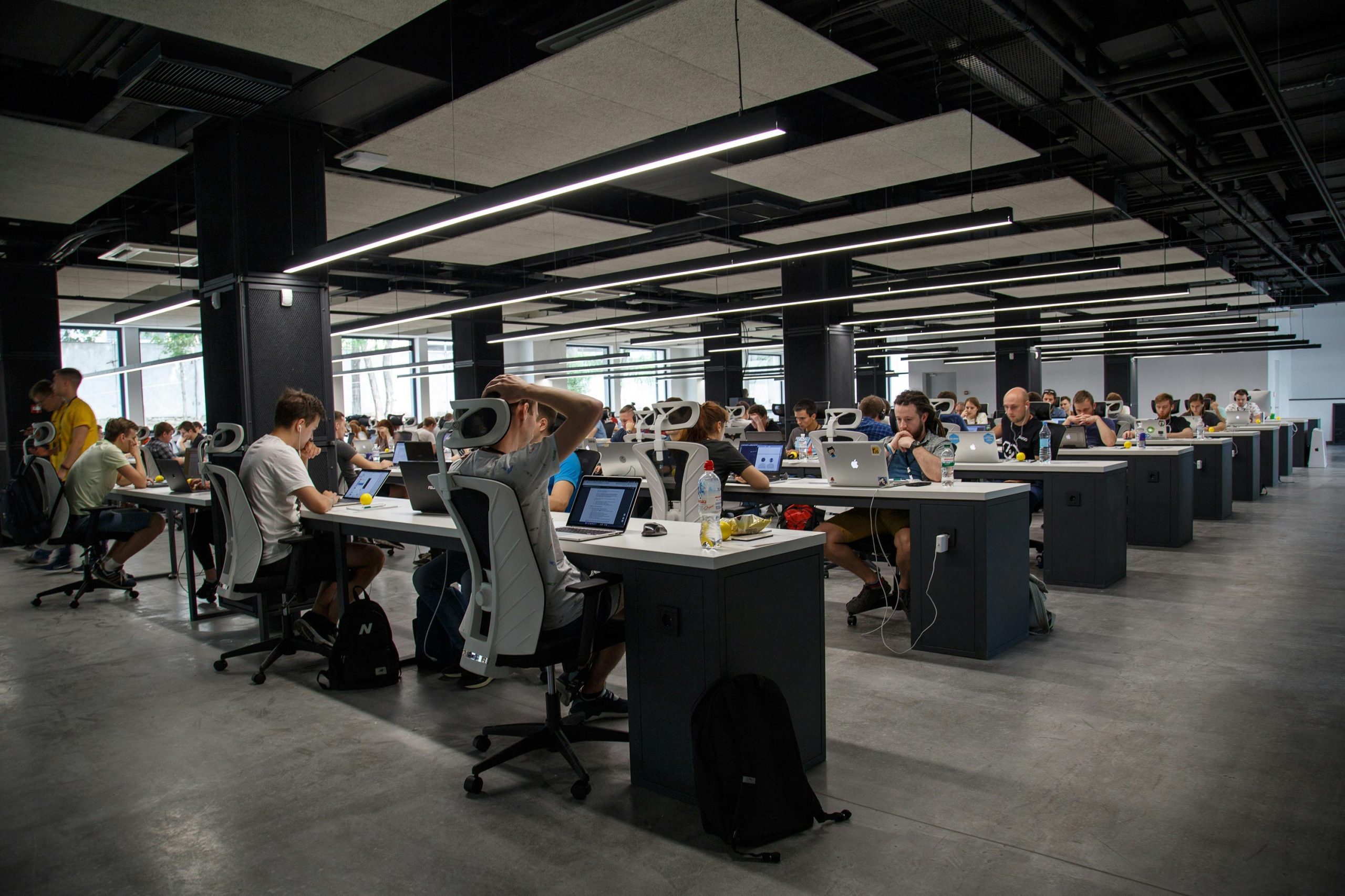 Una oficina espaciosa y moderna con filas de empleados trabajando diligentemente en escritorios largos. Se ven computadoras portátiles y suministros de oficina, creando una atmósfera de concentración.